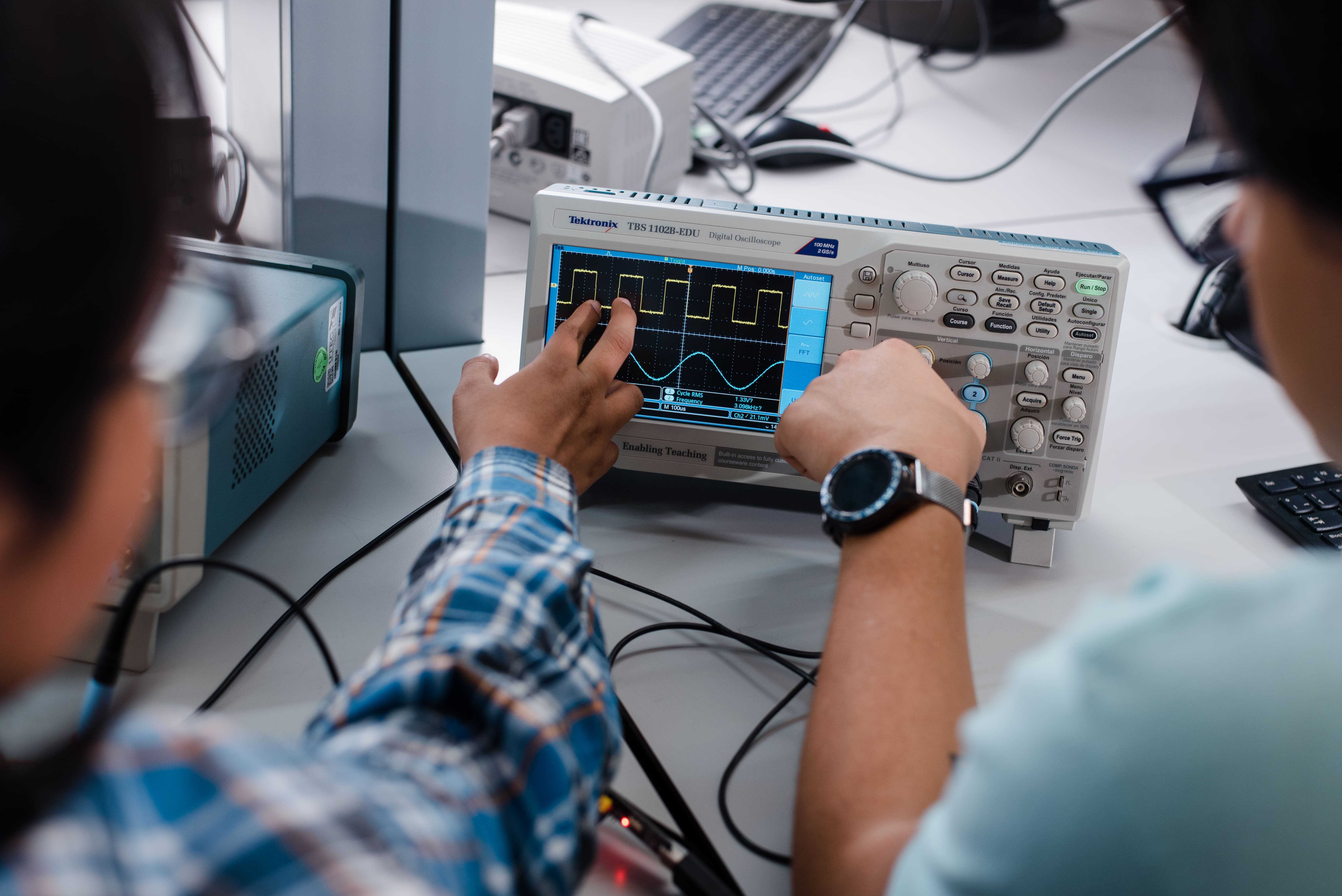 Primer plano de manos de estudiantes operando un osciloscopio digital Tektronix para analizar señales eléctricas durante una práctica de ingeniería en carreras STEM