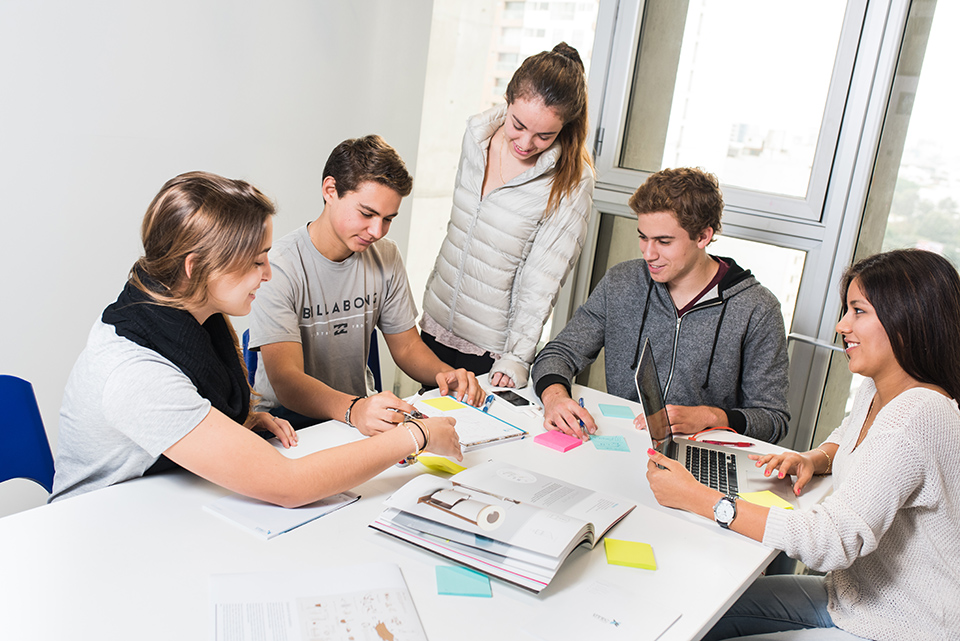 Cinco estudiantes en un espacio de estudio, rodeados de libros y una laptop, revisan materiales educativos para identificar oportunidades de negocio y generar nuevas ideas.